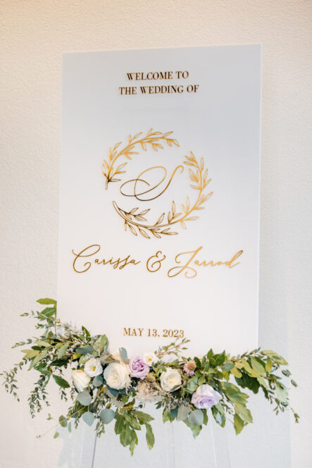 Carissa and Jarrod's White Wedding Welcome Sign with Gold Lettering and Floral Arrangement {{brizy_dc_image_alt imageSrc=