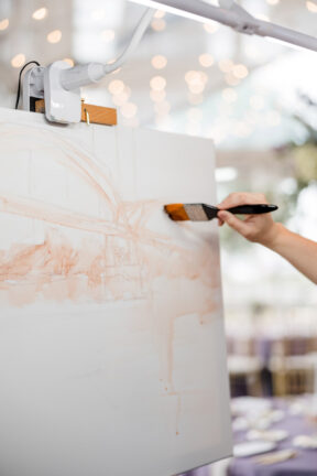 A close-up image of a live wedding painter with her brush to the canvas as she captures the scene {{brizy_dc_image_alt imageSrc=