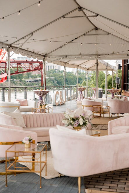 A tented riverfront patio with soft pink lounge furniture for a wedding reception cocktail hour {{brizy_dc_image_alt imageSrc=