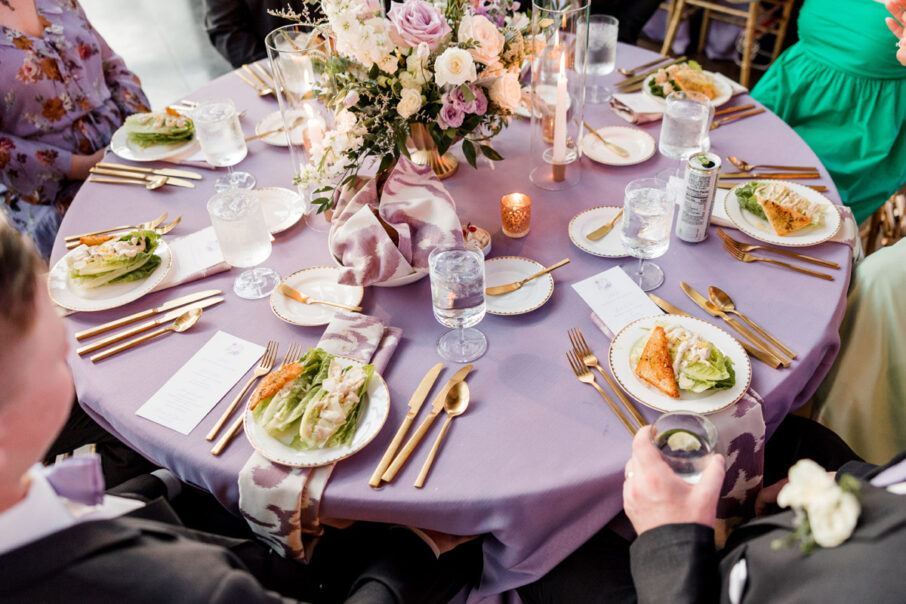 A look down at a reception table with lavender linens and a large floral centerpiece with guests at their seats in front of a plated salad {{brizy_dc_image_alt imageSrc=
