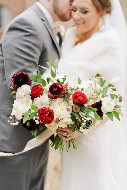 Groom kisses bride's cheek as she smiles in background out of focus with red and white bridal bouquet in foreground for romantic winter wedding at The Bridge Building, downtown Nashville wedding venue {{brizy_dc_image_alt imageSrc=