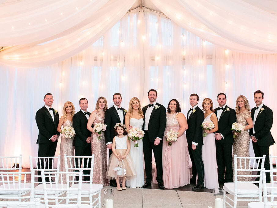 Bride and groom pose with wedding party at ceremony altar before tented wedding ceremony with string lights and white drapery with soft pink lighting for rose gold New Year's Eve wedding at the Bridge Building, a downtown Nashville wedding venue {{brizy_dc_image_alt imageSrc=