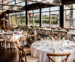 A long farm table with several round table set up for a rehearsal dinner in the Cumberland space of our private event venue, with wooden chairs, white linens, and bright flowers in bud vases for centerpieces {{brizy_dc_image_alt imageSrc=