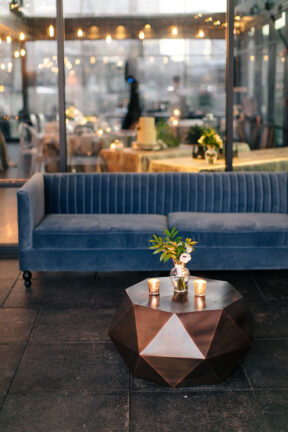 Wedding lounge area setup for cocktail hour with blue velvet sofa and modern bronze coffee table with small bud vase centerpiece for tropical-inspired wedding at the Bridge Building, a downtown Nashville wedding venue