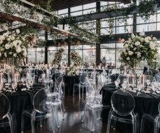 The Cumberland space set up with a grand wedding reception, filled with round tables in black linens, clear acrylic ghost chairs, large white floral centerpieces, a white floral ring hanging at the center, and abundant greenery wrapping the industrial beams of our Nashville wedding venue