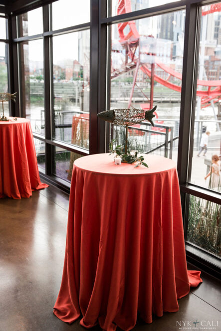 Cocktail tables with red linens and metal fish art centerpiece for nautical themed rehearsal dinner {{brizy_dc_image_alt imageSrc=