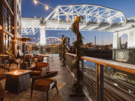 Lounge areas with fire pits set up on the riverfront patio with winter pine decor, overlooking the lit up pedestrian bridge {{brizy_dc_image_alt imageSrc=