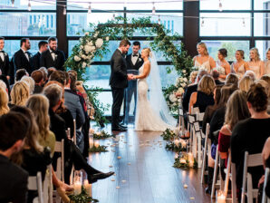 A wedding ceremony in the Dyer space with guests looking on as a bride and groom hold hands at the altar in front of a greenery-wrapped hexagonal arch